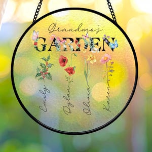May include: A circular hanging suncatcher with a black metal frame and chain. The surface features a yellow and green watercolor background with the text Grandma's GARDEN, colorful floral illustrations, butterflies, and four names: Emily, Dylan, Olivia, and Karen.