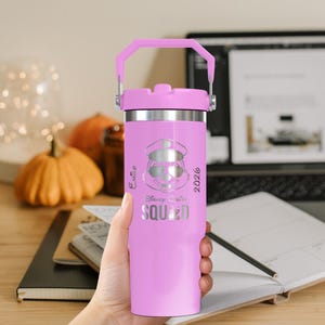 Puede incluir: Un vaso t&eacute;rmico rosa con borde y asa plateados. El vaso presenta un grabado plateado de un personaje de dibujos animados con sombrero y gafas de sol, con las palabras "Disney Cruise Squad 2026".