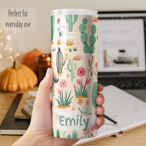 May include: A cylindrical tumbler with a light beige background featuring a repeating pattern of green cacti and pink flowers. The name Emily is printed in dark green letters near the bottom of the tumbler. The item has a clear plastic lid.