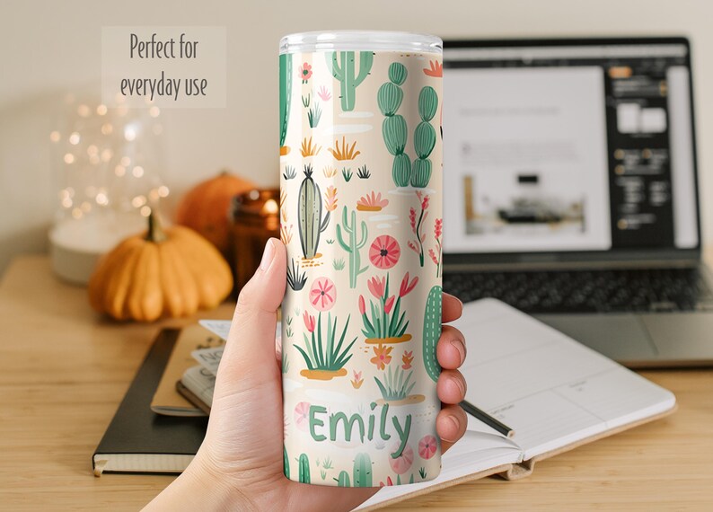 May include: A cylindrical tumbler with a light beige background featuring a repeating pattern of green cacti and pink flowers. The name Emily is printed in dark green letters near the bottom of the tumbler. The item has a clear plastic lid.
