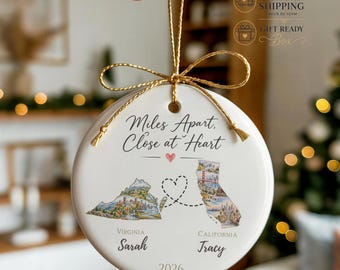 May include: A round white ceramic ornament hanging by a gold string. It features the text Miles Apart, Close at Heart, a small pink heart, maps of Virginia and California connected by a dashed heart line, the names Sarah and Tracy, and 2026.