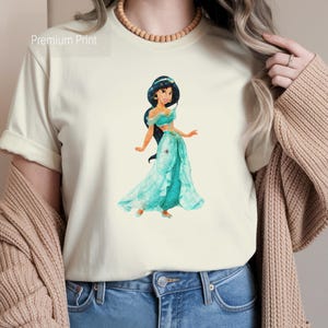 May include: Cream-colored t-shirt featuring a watercolor illustration of a princess in a teal dress. The princess has dark hair and is wearing gold earrings. The text "Premium Print" is in the upper left corner. The person is wearing blue jeans and a tan cardigan.