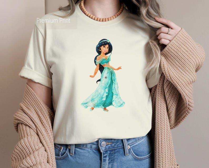 May include: Cream-colored t-shirt featuring a watercolor illustration of a princess in a teal dress. The princess has dark hair and is wearing gold earrings. The text "Premium Print" is in the upper left corner. The person is wearing blue jeans and a tan cardigan.