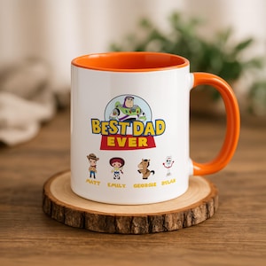 May include: A white ceramic mug with an orange interior and handle. The front features a graphic of Buzz Lightyear above the text "BEST DAD EVER" and four Toy Story characters with the names "MATT," "EMILY," "GEORGIE," and "DYLAN" printed below.