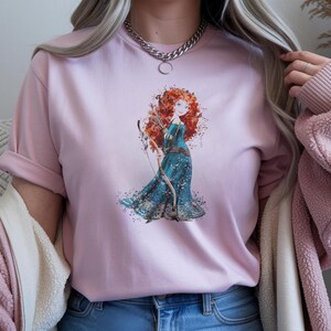 May include: A light pink t-shirt featuring a watercolor illustration of a red-haired woman in a blue dress holding a bow and arrow. The graphic is centered on the shirt. The shirt has rolled-up sleeves.