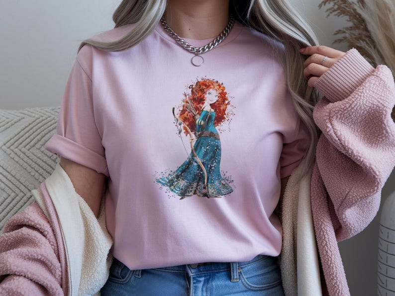May include: A light pink t-shirt featuring a watercolor illustration of a red-haired woman in a blue dress holding a bow and arrow. The graphic is centered on the shirt. The shirt has rolled-up sleeves.