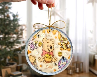 May include: Round Christmas ornament featuring Winnie the Pooh and friends. The ornament has a gold string for hanging and includes the year 2026 and the name "Emily". The design includes honey pots, bees, and flowers.