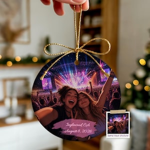 May include: A round ceramic ornament featuring a vibrant concert scene with two people taking a selfie in the foreground. The image includes a pink brushstroke graphic with the text "Inglewood CA August 8, 2026" and a gold string for hanging.