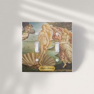 May include: A double light switch cover plate featuring a reproduction of The Birth of Venus painting with soft gold, green, and blue tones. The plate includes two white toggle switches and a gold-colored label with the text Olivia's Bedroom.