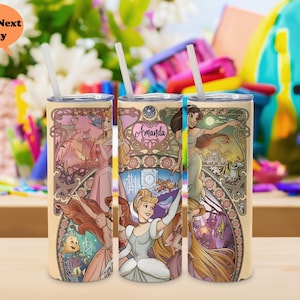 Op de afbeelding: Drie roestvrijstalen bekers met een cr&egrave;mekleurige achtergrond en afbeeldingen van Disney-prinsessen. De prinsessen zijn Assepoester, Rapunzel en Jasmine. De bekers hebben een gouden rand en het woord "Amanda" staat op een van de bekers.