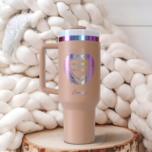 May include: A tan insulated tumbler with a handle, featuring the text "GUARDED HEAVEN" and the name "Emily" in a decorative font. The design includes a shield with iridescent wings. The lid has a metallic rim.
