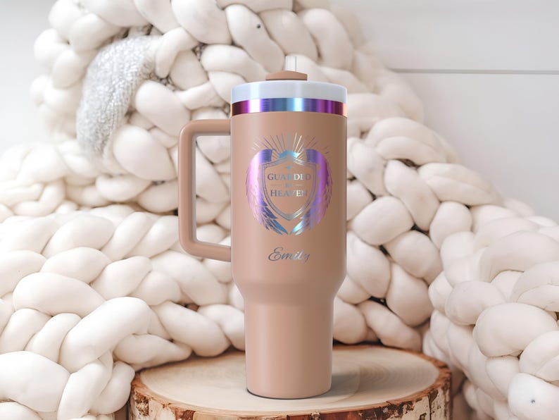 May include: A tan insulated tumbler with a handle, featuring the text "GUARDED HEAVEN" and the name "Emily" in a decorative font. The design includes a shield with iridescent wings. The lid has a metallic rim.