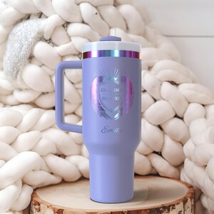 May include: A lavender insulated tumbler with a handle, featuring the text "Guarded Heaven" and the name "Emily". The tumbler has a silver and iridescent top and a matching handle. It is displayed on a wooden surface.