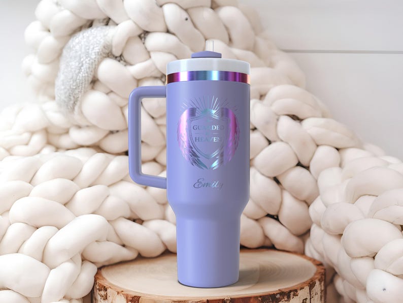 May include: A lavender insulated tumbler with a handle, featuring the text "Guarded Heaven" and the name "Emily". The tumbler has a silver and iridescent top and a matching handle. It is displayed on a wooden surface.