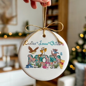 Puede incluir: Un adorno navideño de cerámica blanca con una cuerda dorada. El adorno presenta la palabra "MOM" en un diseño floral, con personajes de dibujos animados. Los nombres "Claire + John + Olivia" están escritos encima de la palabra "MOM".