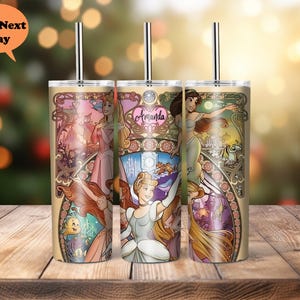 Op de afbeelding: Drie roestvrijstalen bekers met deksels en rietjes. Elke beker heeft een andere Disney-prinses: Ariel, Assepoester en Rapunzel. De bekers hebben een gouden achtergrond met een bloemmotief. De tekst "Amanda" staat op de Assepoester-beker.