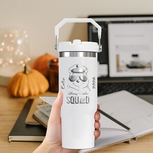 Puede incluir: Un vaso t&eacute;rmico blanco con borde y asa plateados. Presenta un gr&aacute;fico de Winnie the Pooh con gafas de sol y gorra, con el texto "Disney Cruise Squad 2026". El vaso est&aacute; sostenido en una mano.
