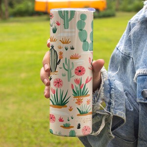 May include: A cylindrical tumbler with a light beige background featuring a repeating pattern of various green cacti and succulents, interspersed with small pink and orange flowers and white horizontal dashes across the entire surface.