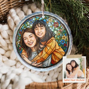 May include: A round, clear glass ornament with a stained-glass style illustration of two smiling people hugging. The ornament has a silver hanging loop and is surrounded by a white knitted blanket and greenery.