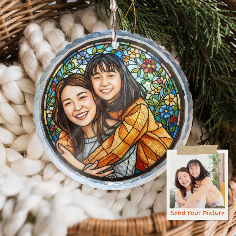 May include: A round, clear glass ornament with a stained-glass style illustration of two smiling people hugging. The ornament has a silver hanging loop and is surrounded by a white knitted blanket and greenery.