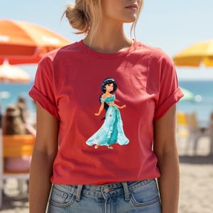 May include: A red t-shirt featuring a graphic of a cartoon princess in a turquoise dress. The shirt has rolled-up sleeves and is paired with blue jeans. The background shows a beach scene with umbrellas and the ocean.
