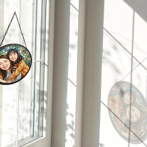 May include: A round stained glass window hanging from a black chain. The window features a portrait of two people, with a colorful floral background. The window is hanging in front of a window.
