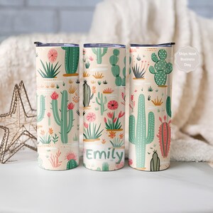 May include: Three cylindrical tumblers with a light beige background featuring a repeating pattern of green and pink cacti and desert plants. The center tumbler includes the name Emily printed in green letters near the bottom of the design.
