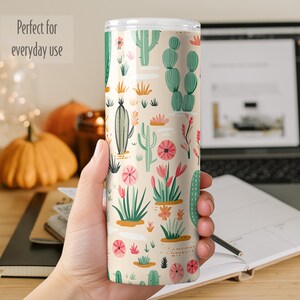 May include: A cylindrical tumbler with a cream-colored background featuring a repeating pattern of various green cacti and pink desert flowers. The tumbler has a clear lid and a smooth finish, displaying a vibrant botanical print across its surface.