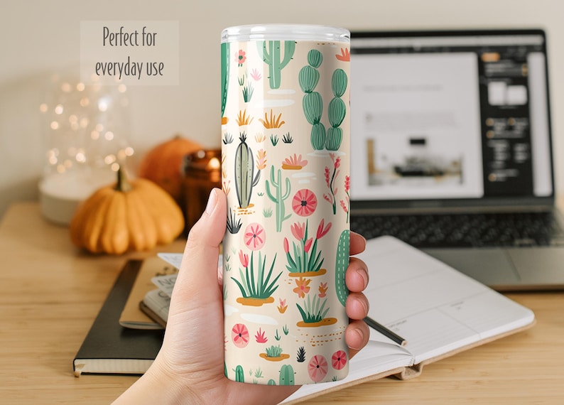 May include: A cylindrical tumbler with a cream-colored background featuring a repeating pattern of various green cacti and pink desert flowers. The tumbler has a clear lid and a smooth finish, displaying a vibrant botanical print across its surface.