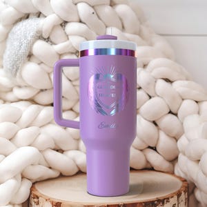 May include: A lavender insulated tumbler with a handle, featuring a white and iridescent design with the text "Guided by Heaven" and the name "Emily". The tumbler has a white and iridescent lid and sits on a wooden surface.