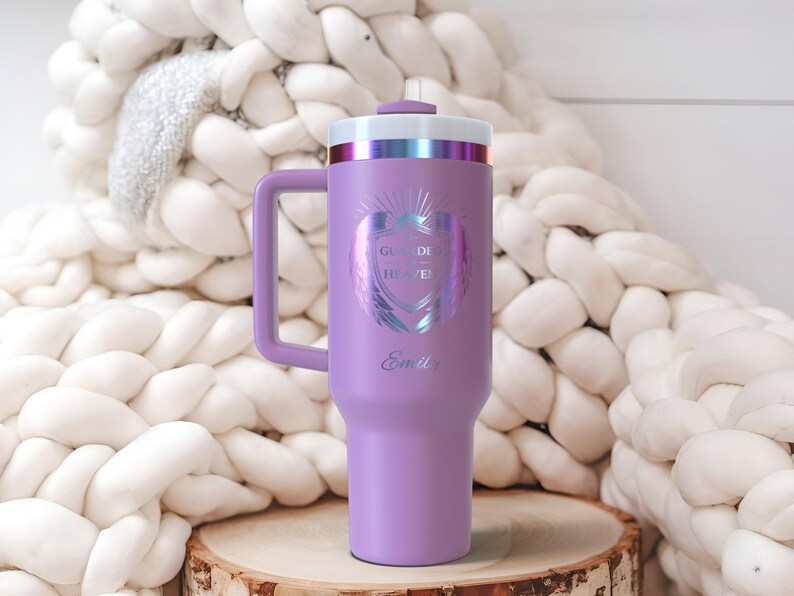 May include: A lavender insulated tumbler with a handle, featuring a white and iridescent design with the text "Guided by Heaven" and the name "Emily". The tumbler has a white and iridescent lid and sits on a wooden surface.