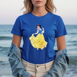 May include: Royal blue t-shirt featuring a watercolor illustration of Snow White in her iconic yellow dress and blue top. The design is centered on the shirt. The model is wearing a denim jacket and khaki pants.