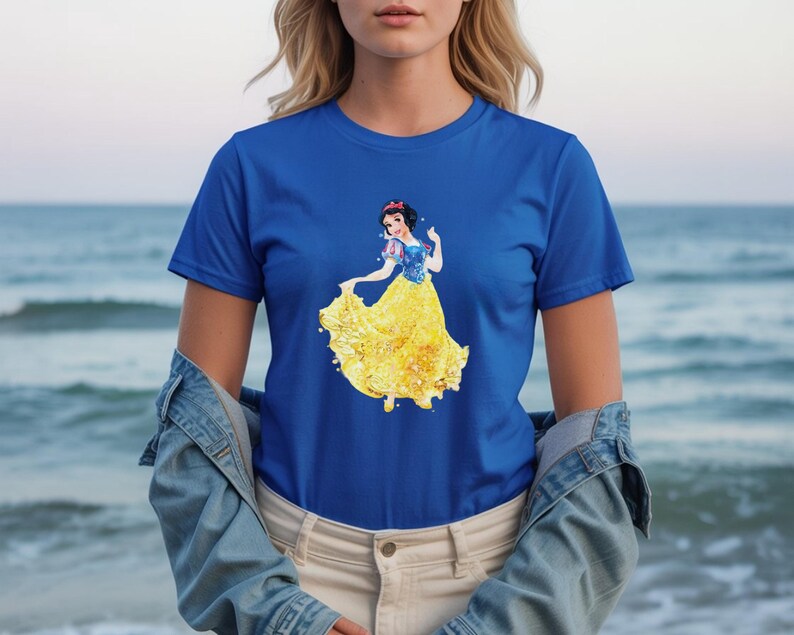 May include: Royal blue t-shirt featuring a watercolor illustration of Snow White in her iconic yellow dress and blue top. The design is centered on the shirt. The model is wearing a denim jacket and khaki pants.