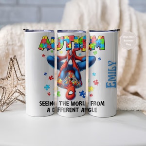 Puede incluir: Vaso blanco con un diseño de piezas de rompecabezas de colores que deletrean "AUTISM" y un gráfico de Spider-Man. El vaso incluye el texto "SEEING THE WORLD FROM A DIFFERENT ANGLE" y el nombre "EMILY" en azul.