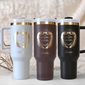 May include: Three insulated tumblers in white, brown, and black, each with a gold emblem that reads "Guarded by Heaven" and the name "Emily." The tumblers have a handle and a gold band around the top.