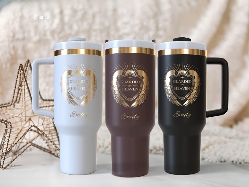 May include: Three insulated tumblers in white, brown, and black, each with a gold emblem that reads "Guarded by Heaven" and the name "Emily." The tumblers have a handle and a gold band around the top.