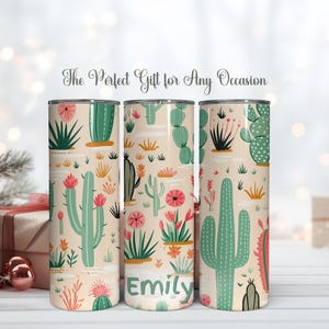May include: Three cylindrical tumblers with a light beige background featuring a repeating pattern of green cacti and pink flowers. The center tumbler displays the name Emily in green text. Each tumbler has a metallic rim at the top and bottom.