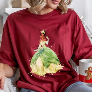 May include: A maroon t-shirt featuring a watercolor illustration of a princess in a green and yellow gown. The princess wears a gold crown. A white mug with a gingerbread man design and a red stripe is held in the hand.