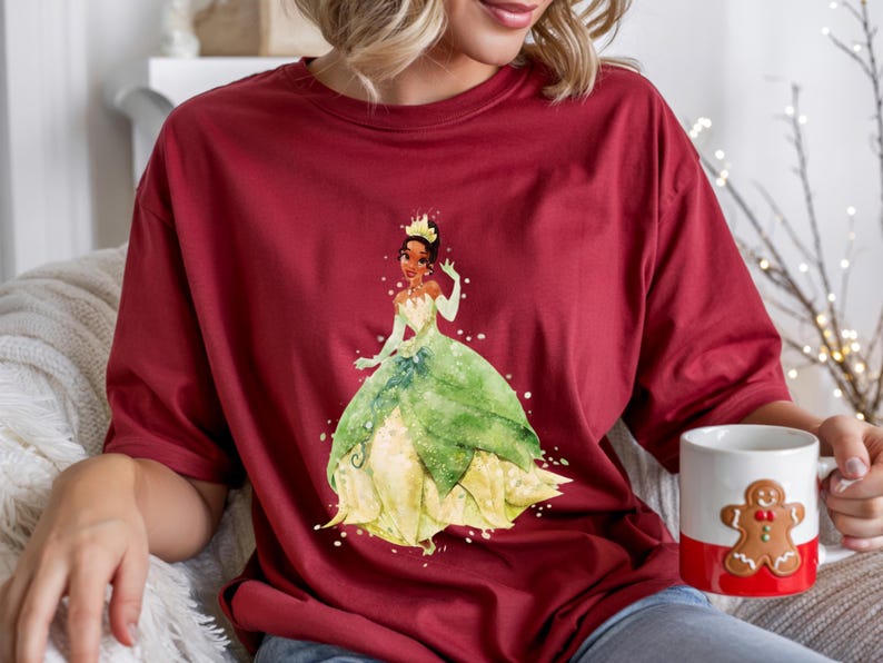 May include: A maroon t-shirt featuring a watercolor illustration of a princess in a green and yellow gown. The princess wears a gold crown. A white mug with a gingerbread man design and a red stripe is held in the hand.