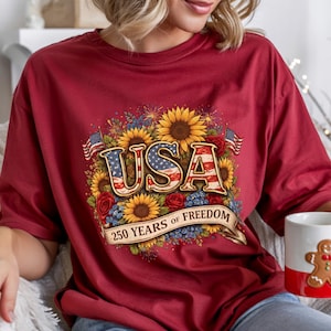Könnte beinhalten: Ein weinrotes T-Shirt mit einem floralen Design mit Sonnenblumen, Rosen und dem Wort "USA" in patriotischen Farben. Das Design enthält den Text "250 YEARS OF FREEDOM" auf einem Banner. Eine weiße Tasse mit einem Lebkuchenmann ist ebenfalls zu sehen.