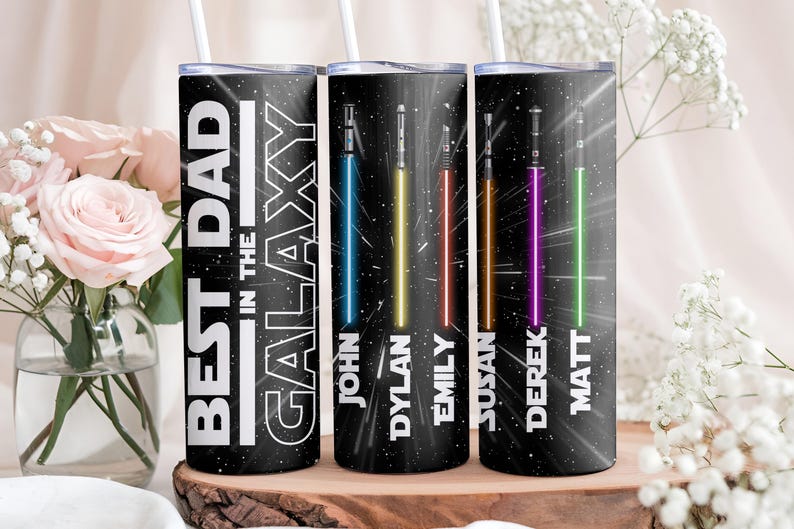 K&ouml;nnte beinhalten: Drei schwarze isolierte Becher mit wei&szlig;en Strohhalmen. Der erste Becher tr&auml;gt in wei&szlig;en Blockbuchstaben die Aufschrift "BEST DAD IN THE GALAXY". Die anderen beiden Becher zeigen farbenfrohe Lichtschwerter und Namen.