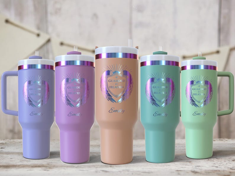 May include: Five insulated tumblers in pastel colors: lavender, purple, peach, teal, and mint green. Each tumbler has a white lid with a silver band, a straw, and a handle. The tumblers are decorated with a shield design and the text "Guarded by Heaven" and "Emily".