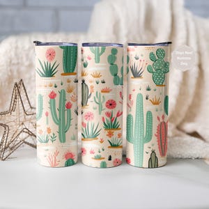 May include: Three tall cylindrical tumblers with lids, featuring a repeating pattern of various green and pink cacti and succulents on a light beige background. The design includes diverse cactus shapes, small flowers, and desert plants.