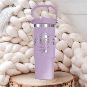 Puede incluir: Un vaso aislado de color lavanda con un borde plateado y un asa a juego. El vaso presenta un gr&aacute;fico de un personaje con gafas de sol y un sombrero, con el texto "Disney Ohana Squad 2026". El vaso est&aacute; sobre una superficie de madera.