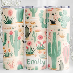 May include: Three cylindrical tumblers with a light peach background featuring a repeating pattern of various green cacti and pink flowers. The center tumbler displays the name Emily in green text. Each tumbler has a clear lid and a smooth finish.