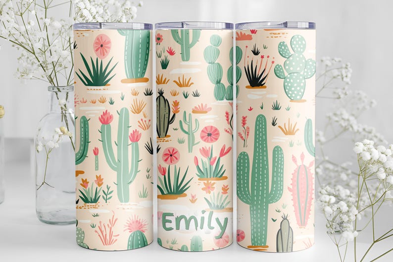 May include: Three cylindrical tumblers with a light peach background featuring a repeating pattern of various green cacti and pink flowers. The center tumbler displays the name Emily in green text. Each tumbler has a clear lid and a smooth finish.