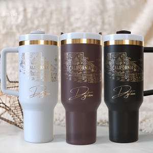 Könnte beinhalten: Drei isolierte Becher in Weiß, Braun und Schwarz, jeweils mit einem goldfarbenen Kalifornien-Karten-Design und dem Namen "Dylan" in eleganter Schrift. Die Becher haben Griffe und goldfarbene Akzente.