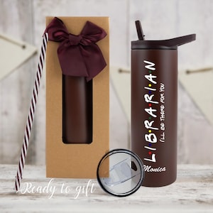Pode incluir: Uma caneca térmica castanha de aço inoxidável com a palavra "LIBRARIAN" e a frase "I'LL BE THERE FOR YOU" impressas em branco. A caneca tem uma tampa castanha escura e uma palhinha. É embalada numa caixa de presente com um laço bordô.