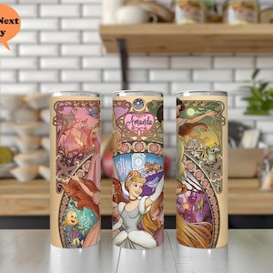 Op de afbeelding: Drie roestvrijstalen bekers met een kleurrijk ontwerp met prinsessen uit Disney's Assepoester. De bekers hebben een gouden rand en een doorzichtig deksel. Het ontwerp bevat een kasteel, een klok en een koets. De tekst "Amanda" staat op een van de bekers.