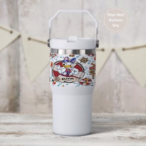 May include: A white insulated tumbler with a handle and a lid. The tumbler features a wrap-around design with a cartoon Donald Duck illustration, nautical elements, and the name "Olivia". The tumbler is designed to keep beverages cold or hot.